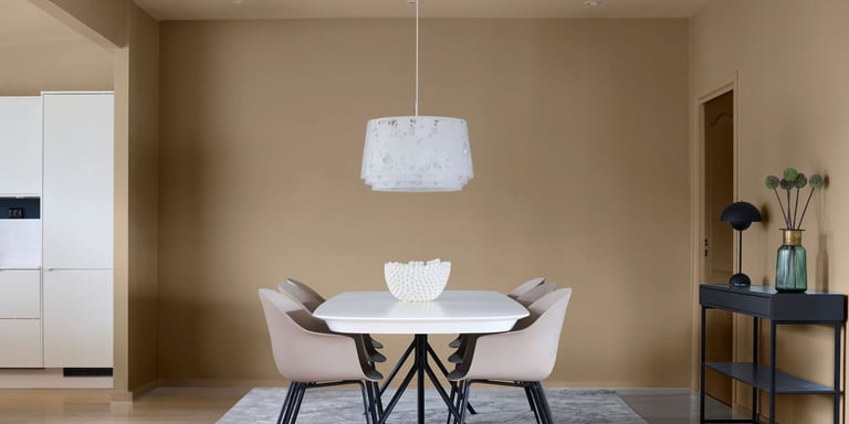 Spisebord med stoler og en hvit lampe over seg. Spisestuen står på et lysegrått gulvteppe. Veggene er malt i en varm oransje-beige farge.