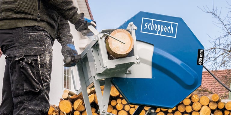 Person bruker Scheppach vedkløyver med 405 mm blad til å kappe vedkubbe foran stor vedstabel.