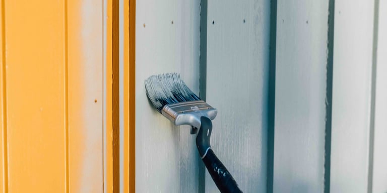 En malingspensel som maler grått på et gult hus.