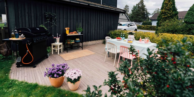 En stor uteplass med terrasse med grill, dekket bord, fargerike stoler i grønne omgivelser foran et svart hus