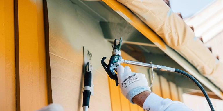 Person som bruker malingssprøyte på et gult hus.