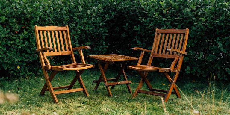 To stoler og et bord av tre står ute i en grønn og frodig hage.