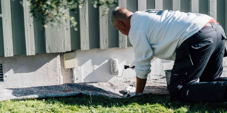 Mann i hvit genser som maler grunnmuren på huset hvit